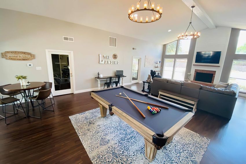 A spacious living area featuring a pool table in the foreground, surrounded by a decorative rug. In the background, there is a cozy seating arrangement with a large sofa, a wall-mounted TV, and large windows allowing natural light to fill the space. A dining table with chairs is visible to the left. Modern decor and lighting contribute to a warm, inviting atmosphere.