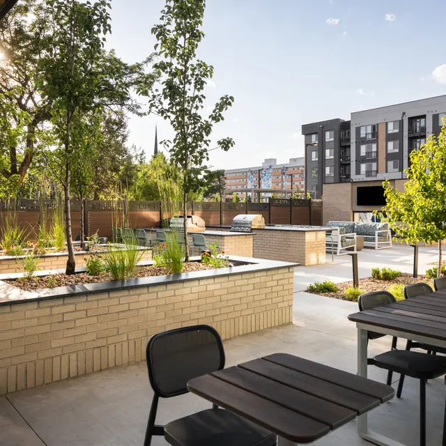 A landscaped communal outdoor area with trees, planters, and picnic-style seating. In the background, modern apartment buildings can be seen under a clear blue sky.