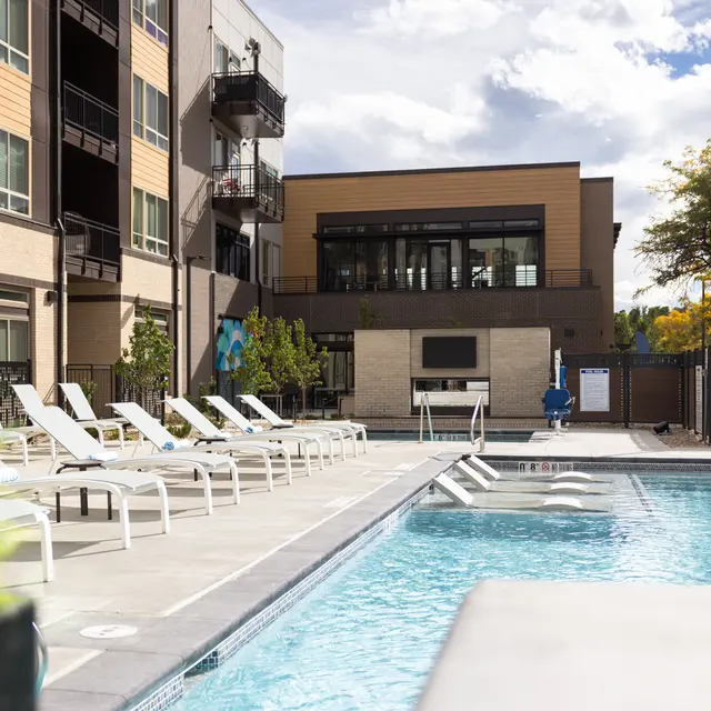 A well-maintained outdoor pool area surrounded by lounge chairs and an apartment building in the background.