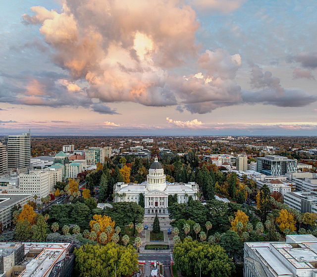 Breathtaking aerial view of a vibrant cityscape highlighting lush greenery, a majestic classical building, and dramatic sunset clouds.