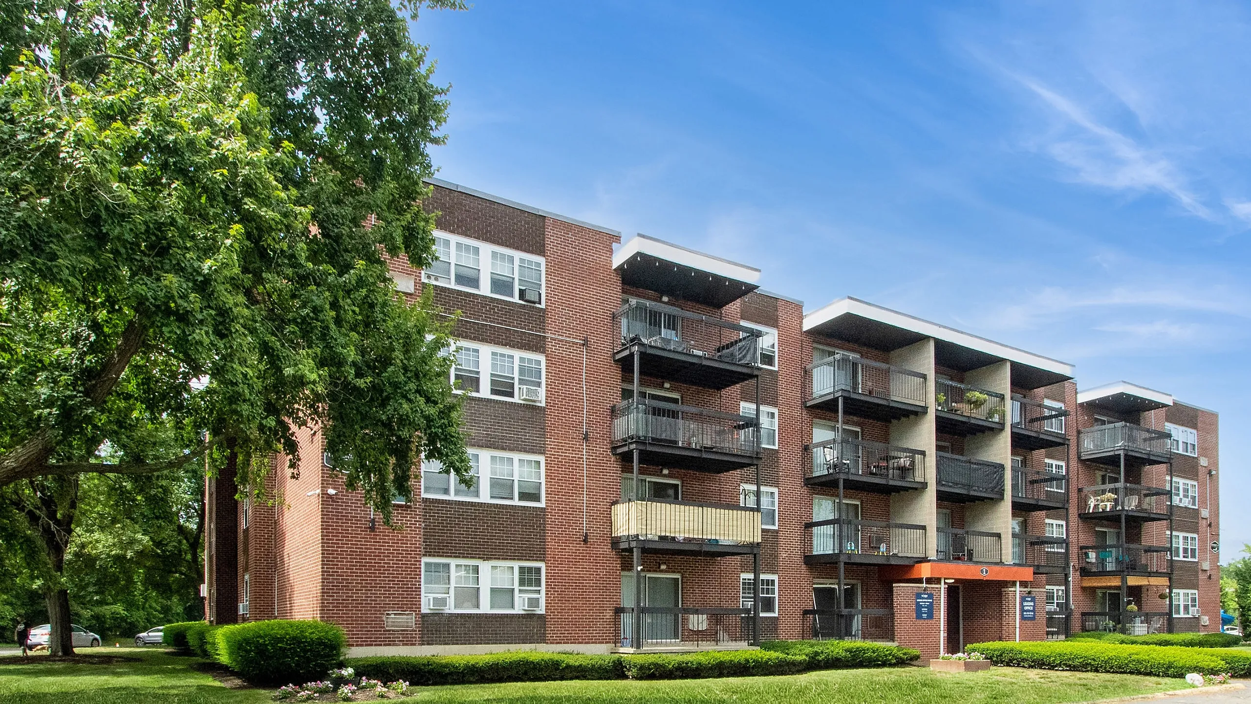 Modern brick apartment building with spacious balconies surrounded by lush greenery and a clear blue sky.