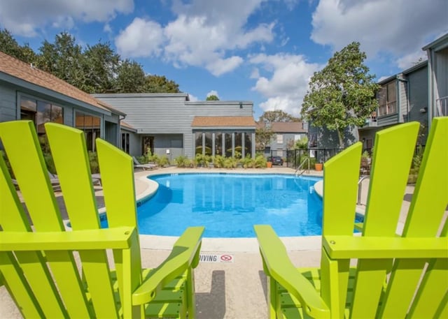 Bright green Adirondack chairs face a sparkling blue swimming pool, with apartment buildings and trees in the background under a cloudy sky.
