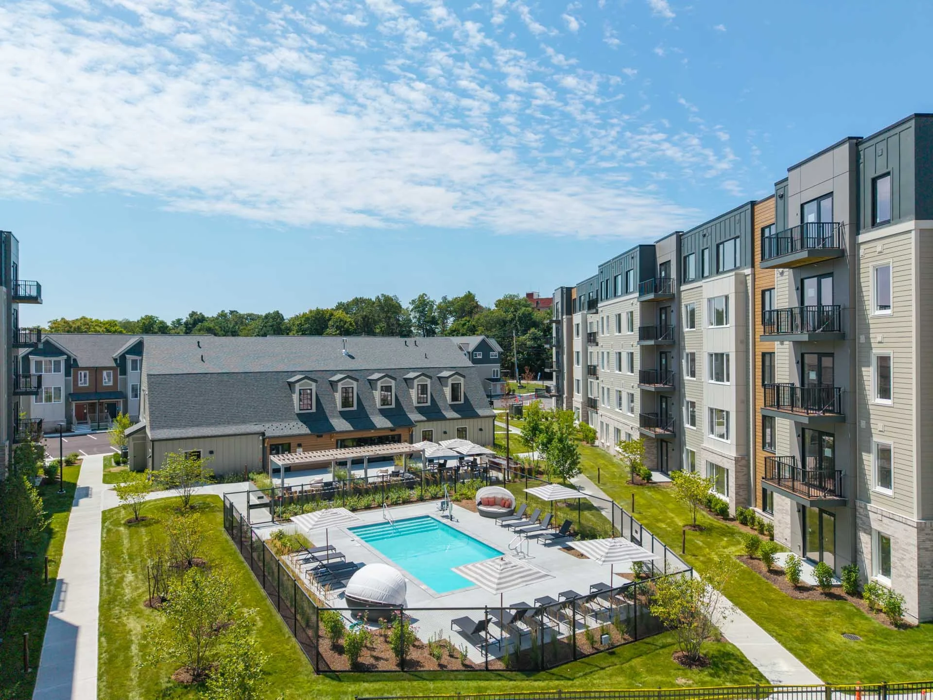Apartment complex with a swimming pool, surrounded by grassy areas and trees, with modern multi-story apartment buildings and neighboring houses under a partly cloudy sky.