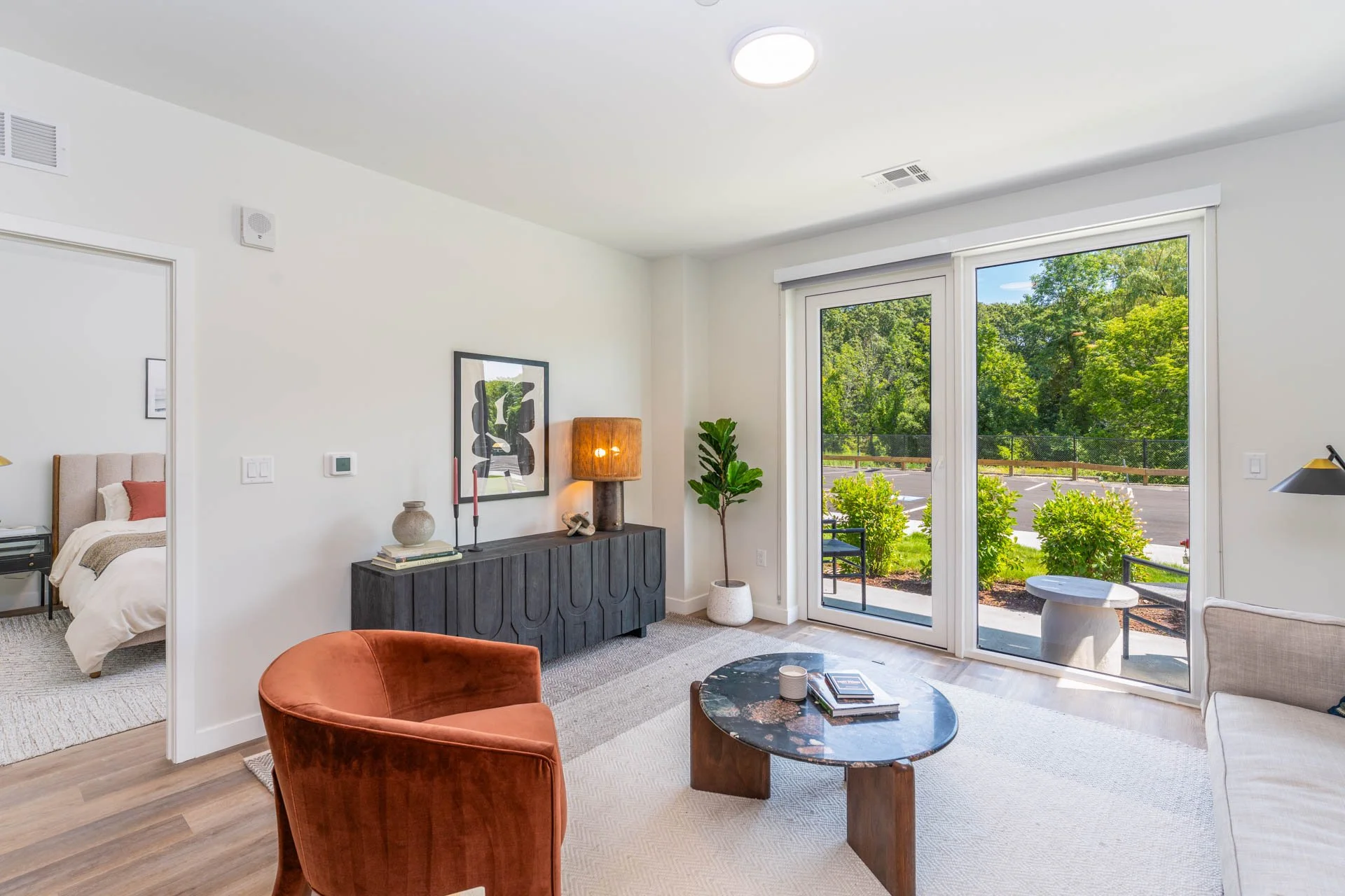 Model apartment living room with sliding glass door leading outside, view of trees and parking lot, beige sofa, rust-colored armchair, black sideboard, potted plants, coffee table, decorative items, bedroom visible through doorway.