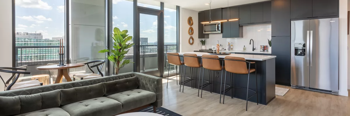 Living room with large windows and wood-style flooring that opens to a kitchen with black cabinets and an island with bar stools.