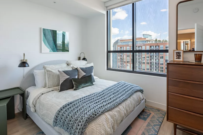 Bright bedroom with a large window, a gray upholstered bed, patterned bedding, and a mid-century modern dresser.