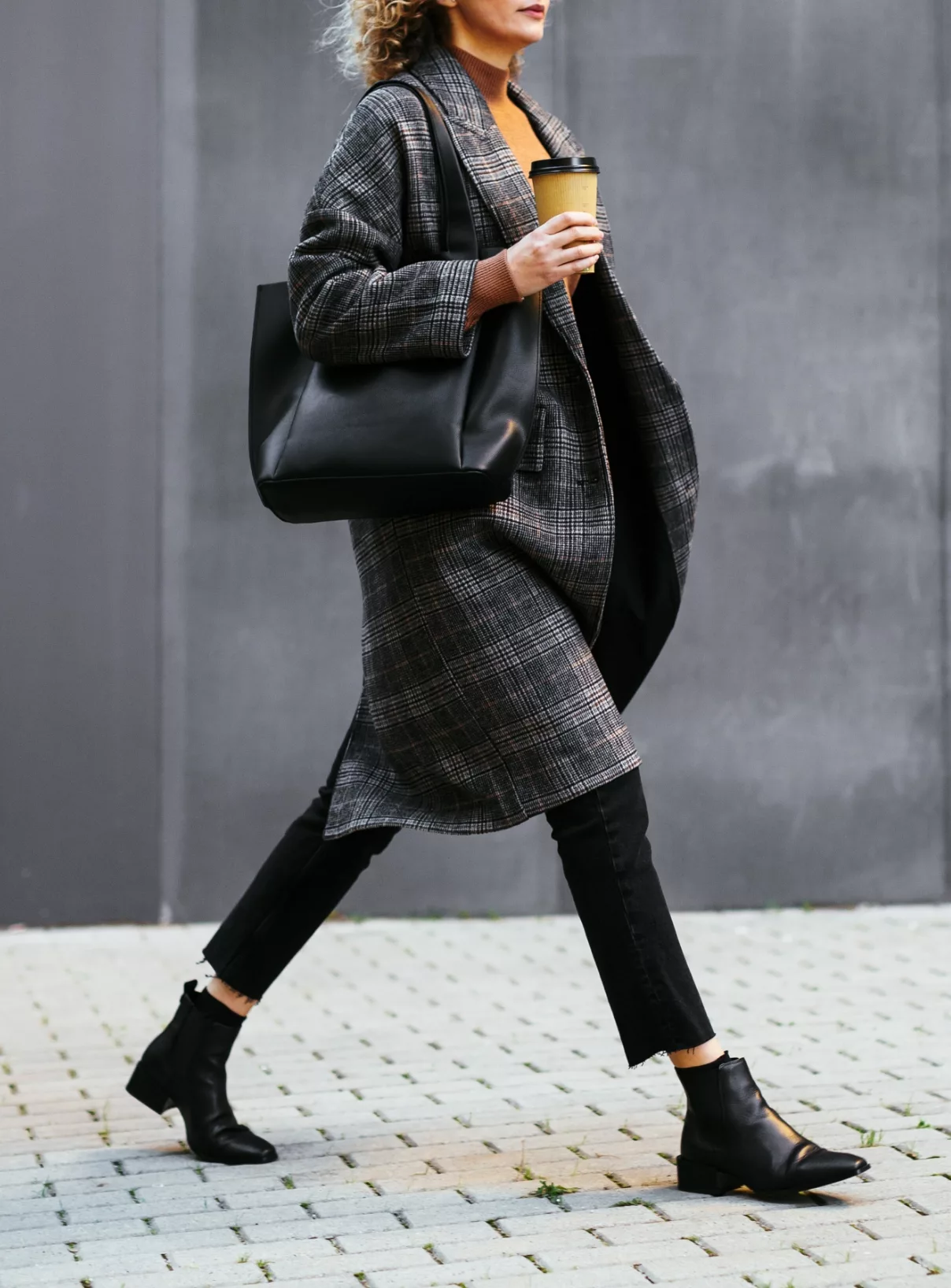 Person walking with a coffee cup, wearing a plaid coat, black pants, and black ankle boots, carrying a large black bag against a grey wall.