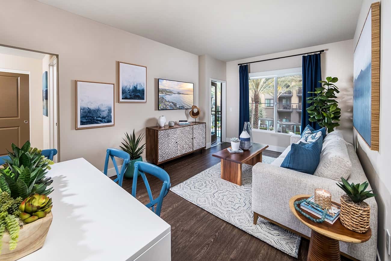 Colony at the Lakes - Plan B1 - Living room with modern furniture and a large window showing palm trees outside.