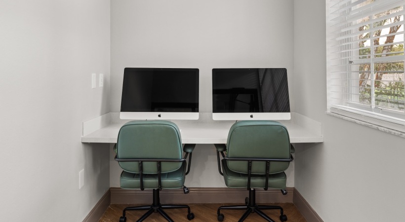 Two computer monitors are sitting on a desk with green chairs.