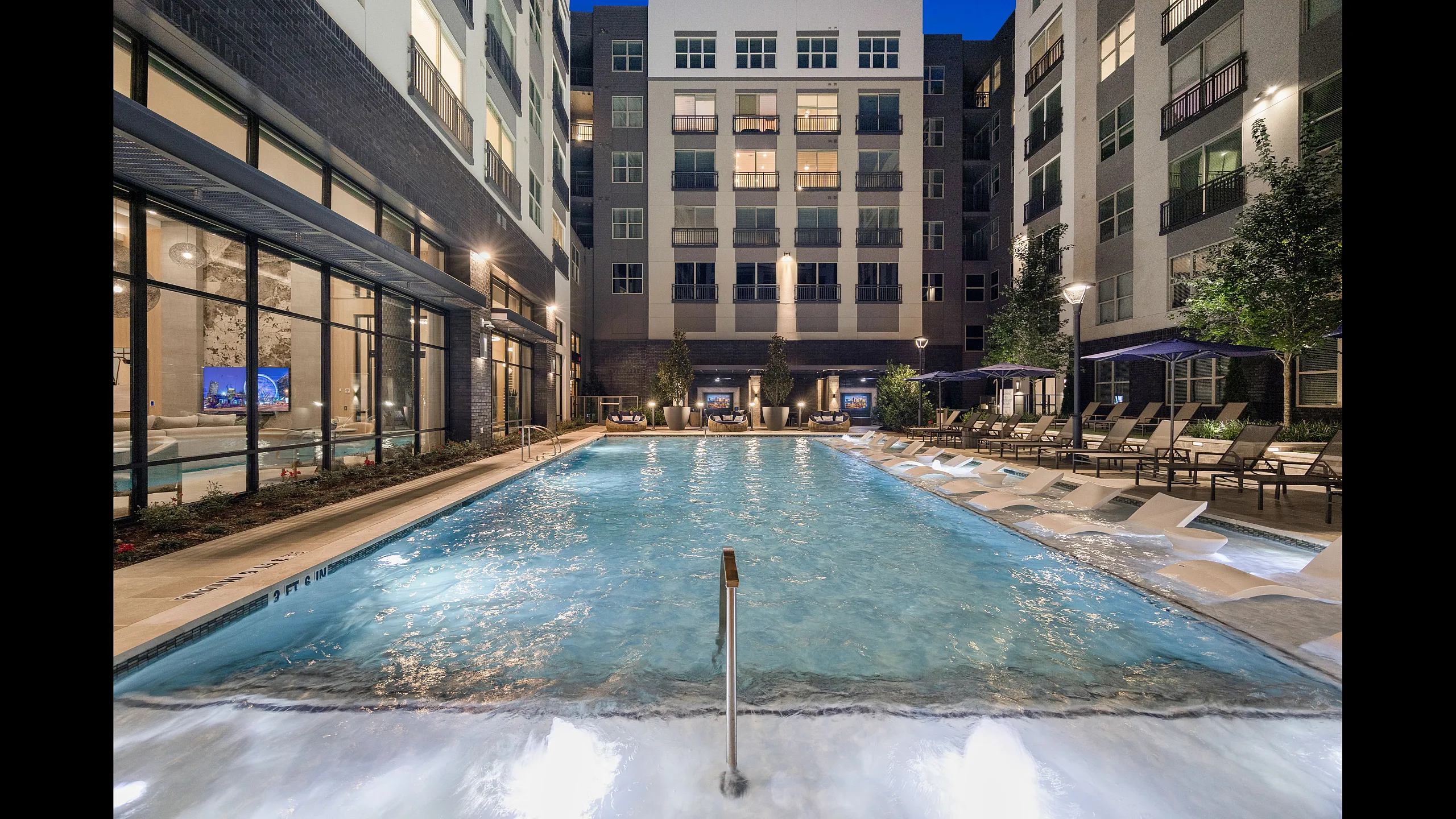 Luxurious apartment complex featuring a sparkling pool with stylish lounge chairs, modern architecture, and elegant outdoor lighting at dusk.
