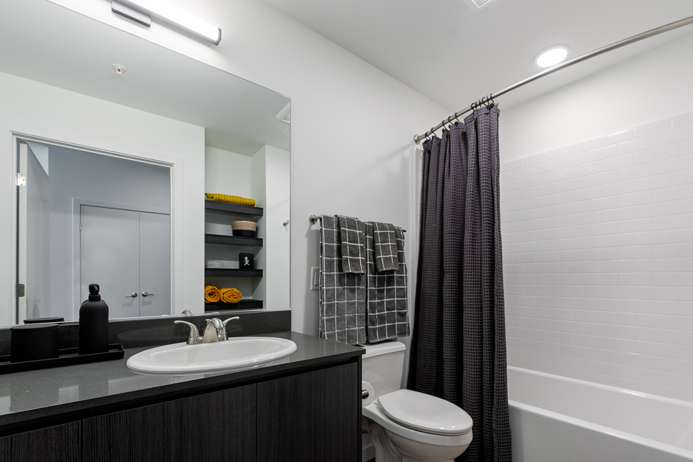 Modern bathroom with dark wood vanity, white sink, and black countertop. Gray towels hang above the toilet, and a dark gray shower curtain covers a white-tiled bathtub. Open shelving holds yellow towels and decor.