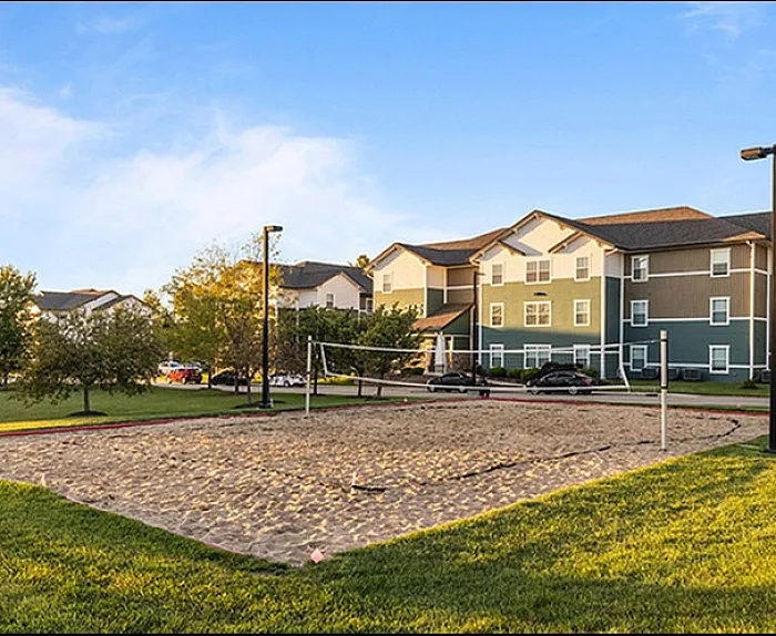 Charming apartment complex with spacious outdoor area featuring a sand volleyball court set against a backdrop of lush greenery and modern architecture.