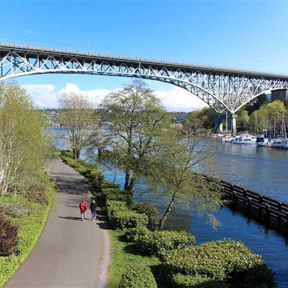 A pedestrian pathway runs alongside a river under a bridge.