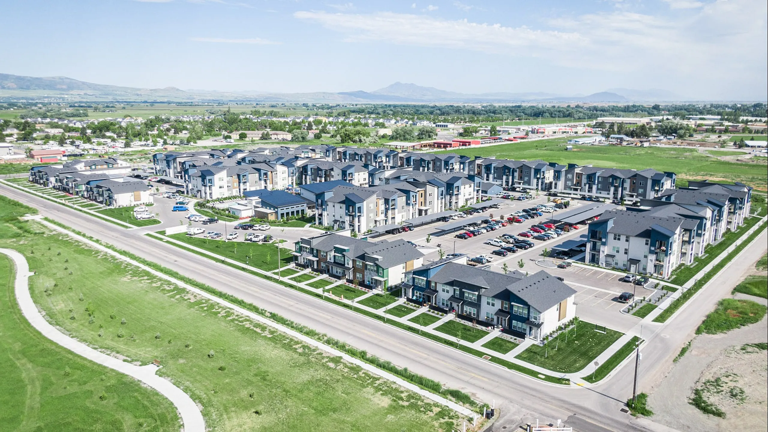 Scenic aerial view of a modern apartment complex with ample parking and lush greenery, surrounded by picturesque landscapes and mountain views.