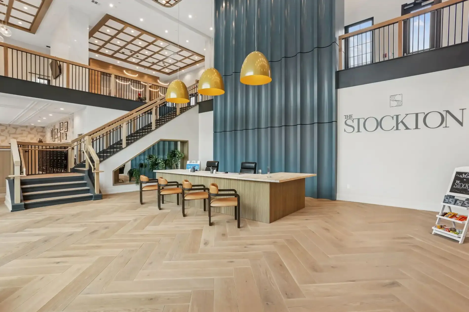 A bright, spacious lobby with a wooden reception desk, gold pendant lights, a grand staircase, and 