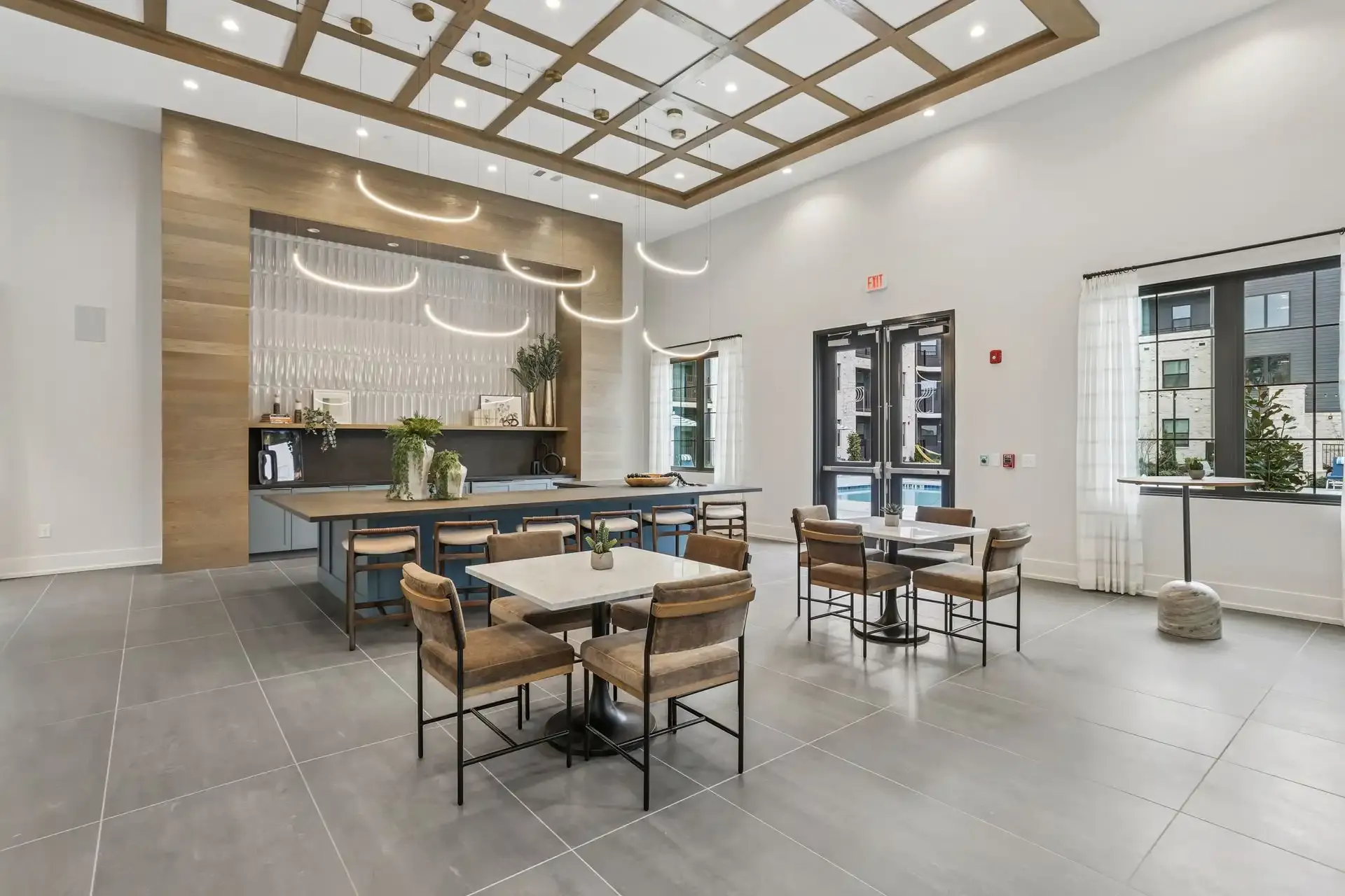 A modern, airy apartment building lounge with tile floors, a coffered ceiling, a central island, and dining tables.