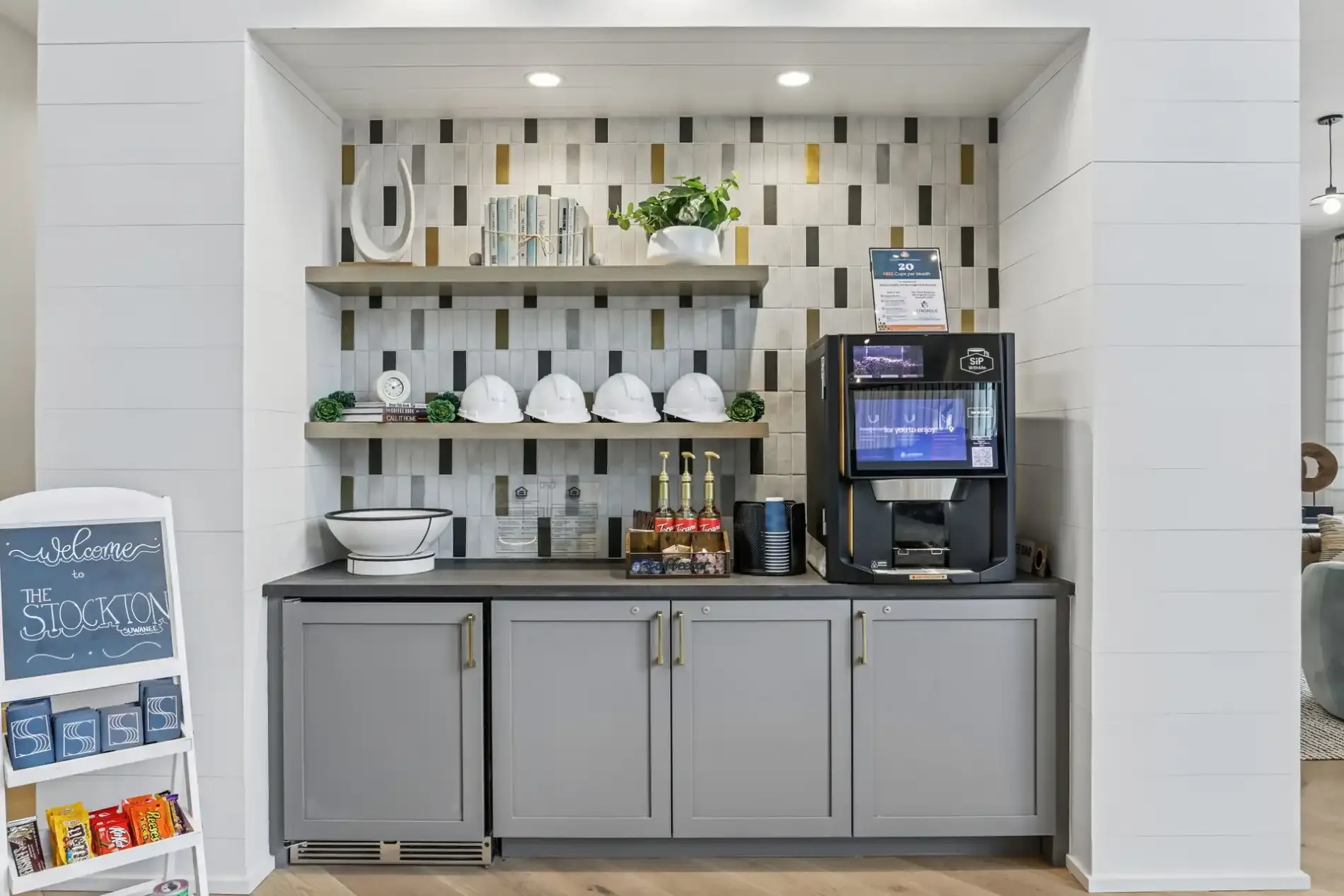 A coffee station in a modern breakroom with a beverage machine, shelving, white hard hats, and gray cabinetry.