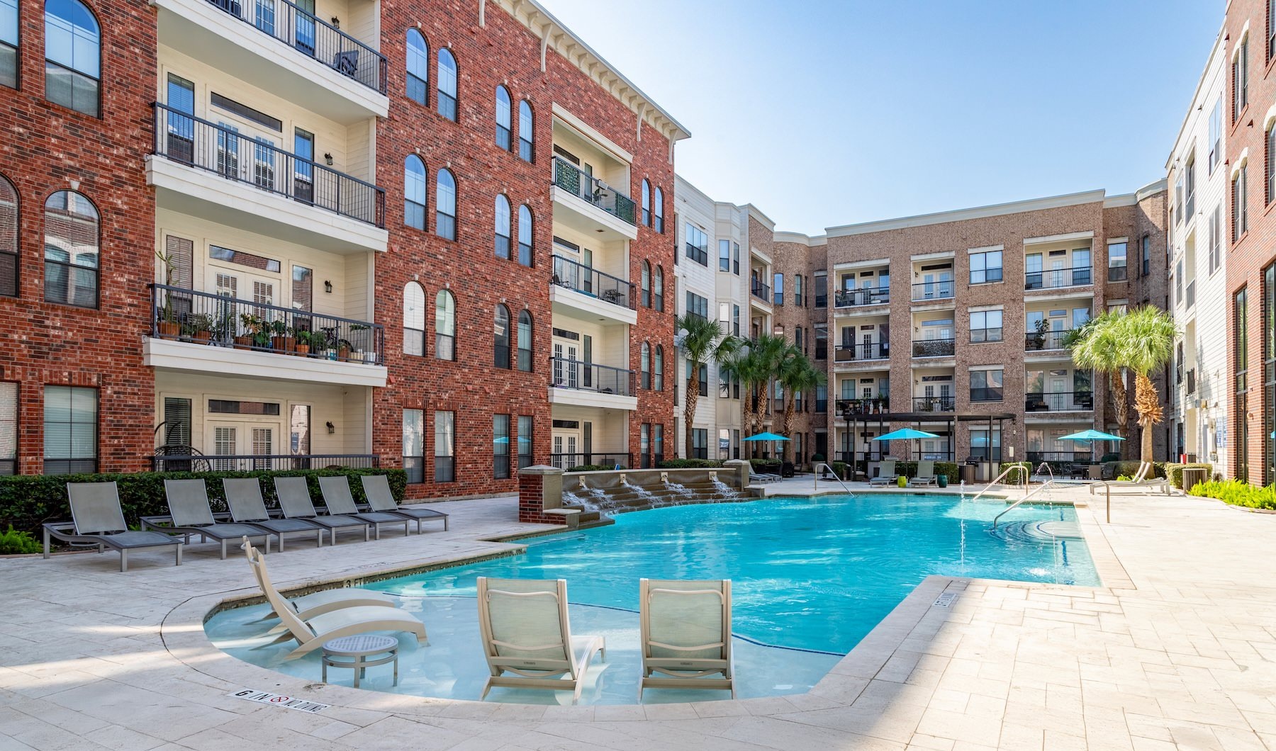 Tanglewood, Houston, TX, Apartments - Forum on San Felipe - Resort-Style Pool with Rippling Fountain Wall with Multiple Lounge Seats, Tanning Ledge, and the Apartment View.
