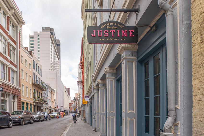 A street scene featuring a restaurant sign for "Justine" located on a building. The sign hangs above a narrow street lined with parked cars and historic architecture, showcasing a mix of modern and traditional buildings in the background under a cloudy sky.