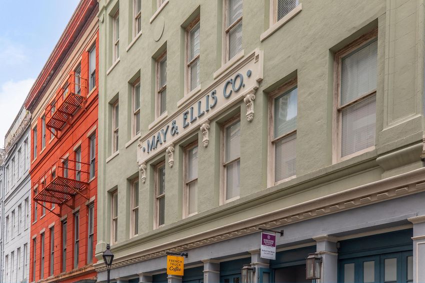 Facade of a historic building with the sign "MAY & ELLIS CO." prominently displayed. The structure features a light green color, with decorative detailing around the windows and door. Nearby, a contrasting red building with fire escapes adds to the urban landscape.