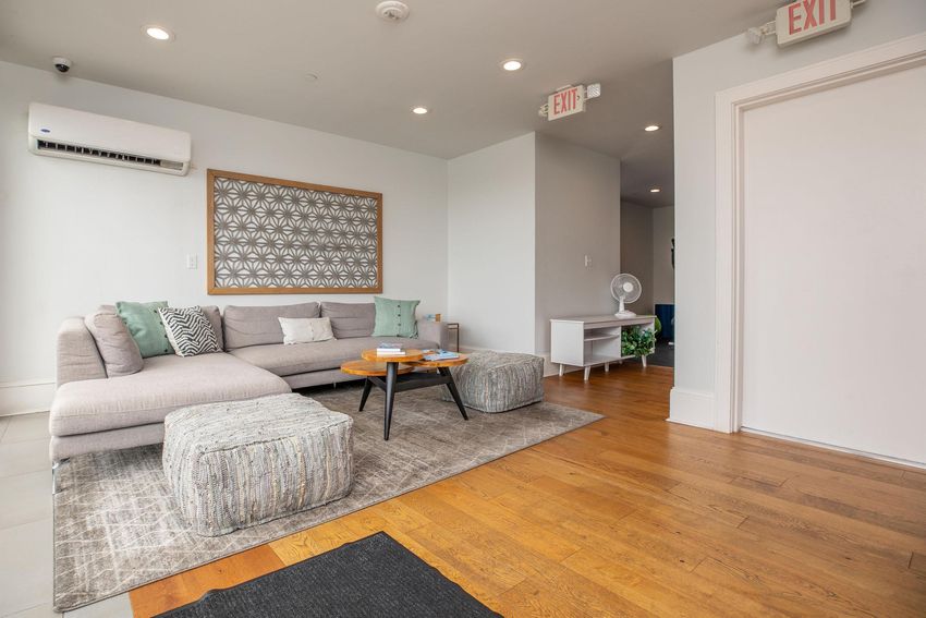 A modern living room with a light gray sectional sofa and decorative pillows. There are two textured ottomans in front of a wooden coffee table. The floors are brown wooden planks, and the walls are painted light gray. A patterned wall decoration is visible, along with an exit door and a fan in the background.