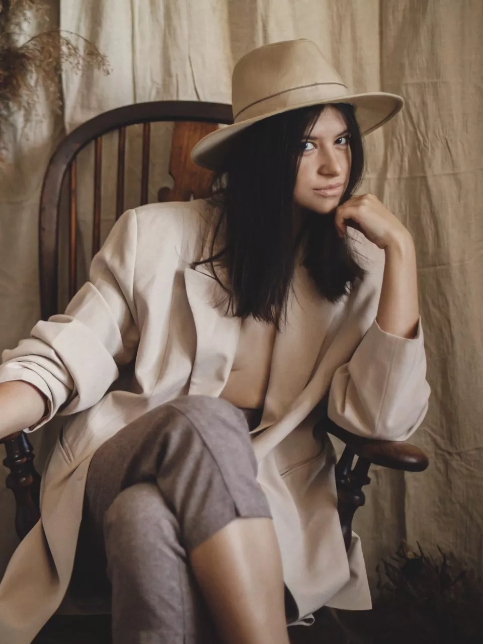 Stylish fashionable woman in beige suit and hat posing on wooden chair in bohemian style room with dried grass and flowers in vase. Young attractive female sitting in atmospheric boho interior