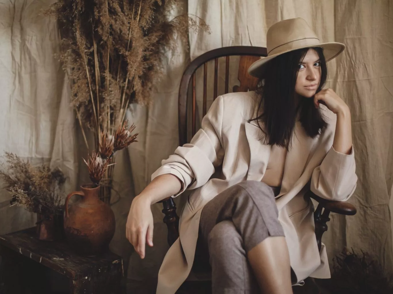 Stylish fashionable woman in beige suit and hat posing on wooden chair in bohemian style room with dried grass and flowers in vase. Young attractive female sitting in atmospheric boho interior