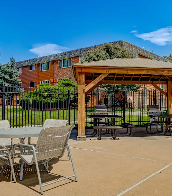 A wooden pavilion with a table and chairs is in the foreground of a sunny outdoor area.