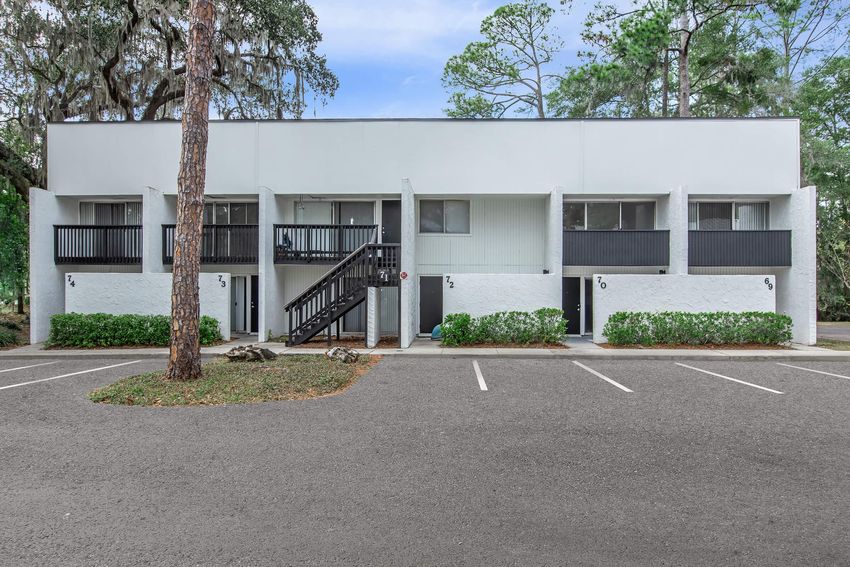 A two-story building with several units, featuring a black staircase in the center. Each unit has a private entrance with small balconies. The surrounding area is landscaped with green shrubs and trees, and there's a gray parking lot in front of the building. The sky is clear and blue.