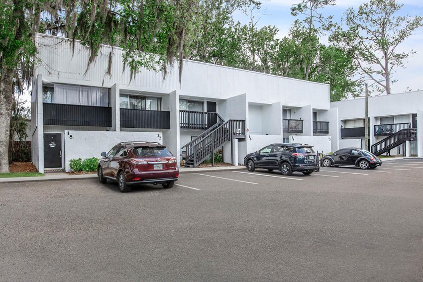 Row of white motel-style buildings with black railings, featuring several parked cars in front. The scene is surrounded by trees, with a clear sky above. The parking lot has designated spaces, and the buildings have a clean, modern design.