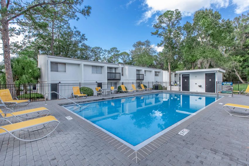 A sparkling blue swimming pool surrounded by gray paving stones, with yellow lounge chairs lining the edges. In the background, there are several white buildings with large windows set among green trees and a clear blue sky, creating a serene outdoor atmosphere.
