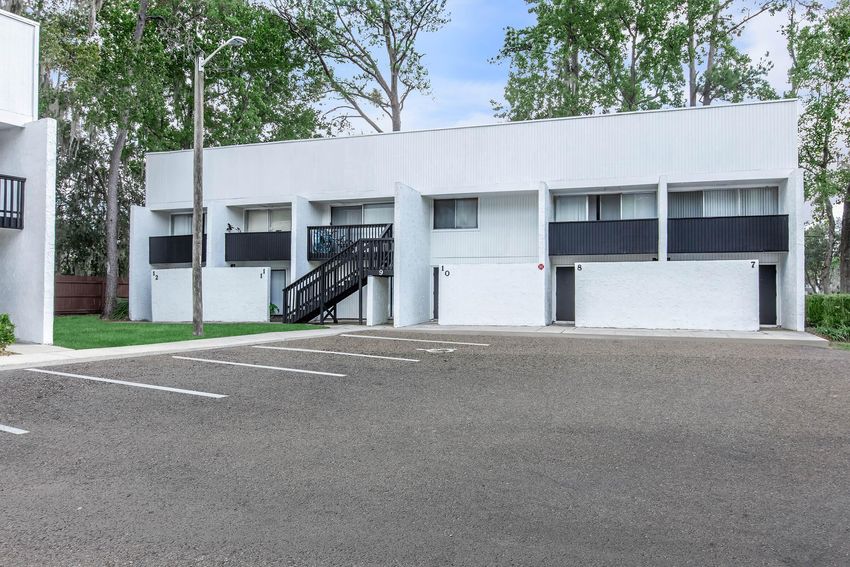 Two-story residential building with white walls and black accents, featuring several doors and windows. A set of stairs leads to the upper level, and there is a parking lot with empty spaces in front. Surrounding greenery includes tall trees, contributing to a peaceful neighborhood atmosphere.