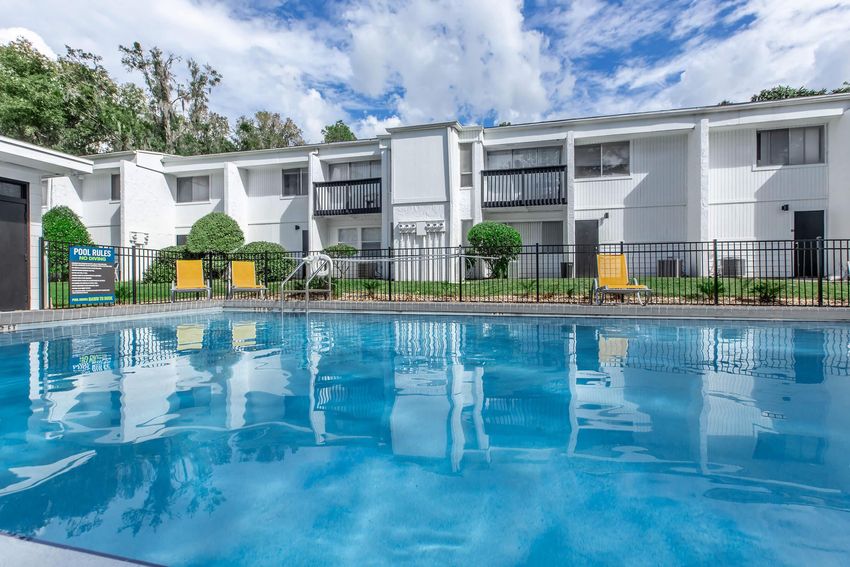 A swimming pool surrounded by lush greenery and a well-maintained landscape. The vibrant blue water reflects the sky and nearby buildings. Yellow lounge chairs are positioned near the pool, and a sign with pool rules is visible in the background, enhancing the inviting atmosphere of the outdoor area.