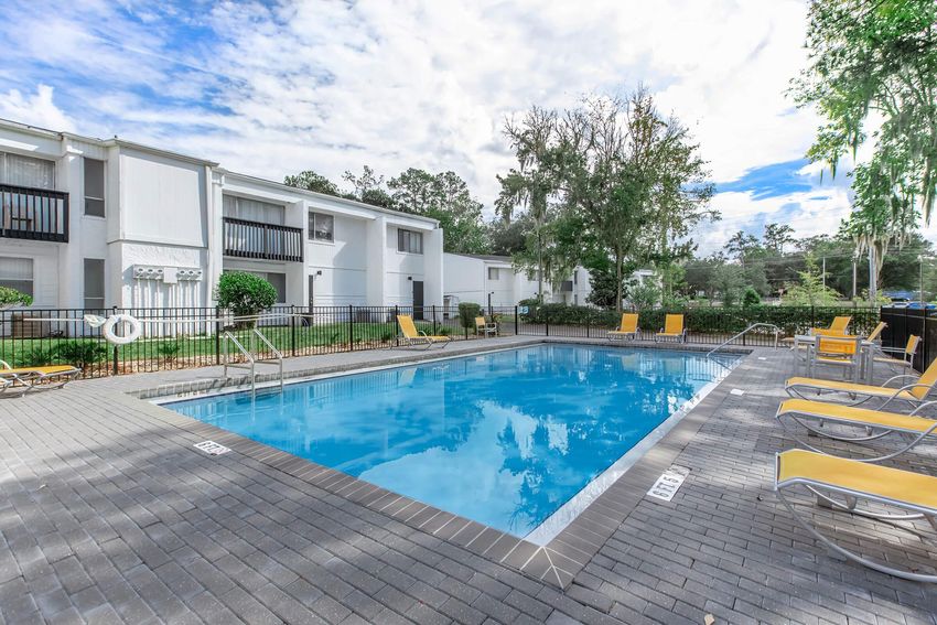 A clean, inviting swimming pool surrounded by lounge chairs, set in a residential area. The pool area features well-maintained landscaping with trees in the background and a clear blue sky. Nearby, there are white residential buildings with balconies visible.