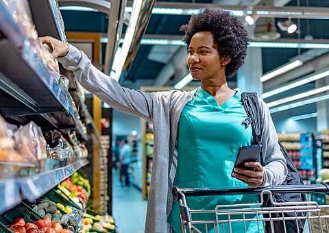 Woman in green scrubs shopping at a brightly lit supermarket, reaching for items on a shelf with a cart filled with fresh produce.