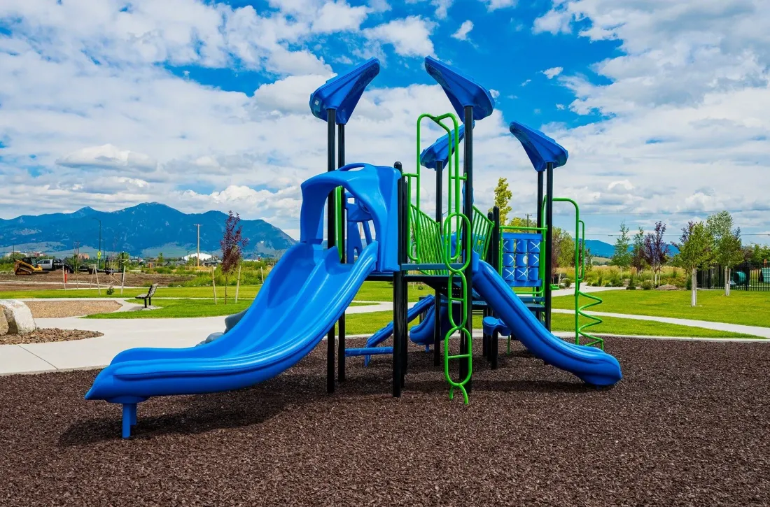 Blue playground climbing structure and slides on a gravel surface under a bright sky with mountains in the background.