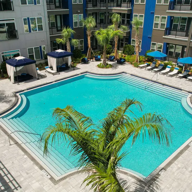 Aerial view of a modern apartment complex pool area featuring a large swimming pool surrounded by palm trees and lounge chairs.