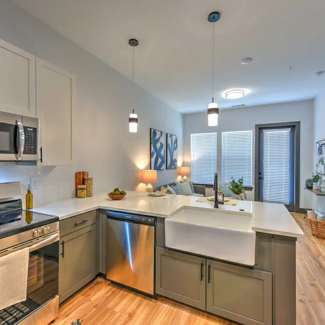 A modern kitchen featuring stainless steel appliances, white cabinetry, and a farmhouse sink. The area has wooden flooring and warm lighting with an adjoining living space visible.