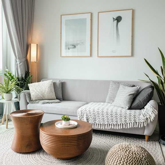 A living room with a grey couch, a brown vase, a round knitted ottoman, and two framed pictures on the wall.