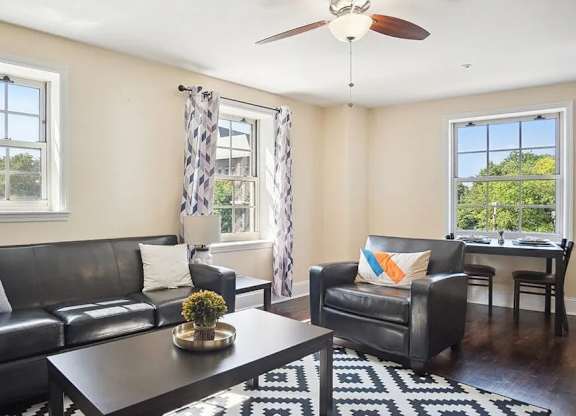 A black leather couch sits in a living room with a ceiling fan and a window with a view of trees.