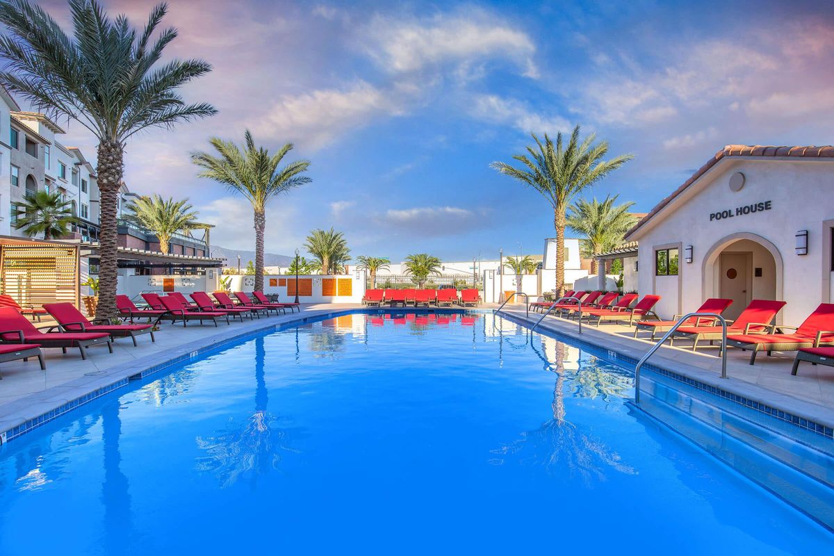 A serene swimming pool surrounded by vibrant palm trees and red lounge chairs. The clear blue water reflects the sky, while a pool house is visible in the background, adding to the inviting atmosphere. This outdoor space is perfect for relaxation and leisure.
