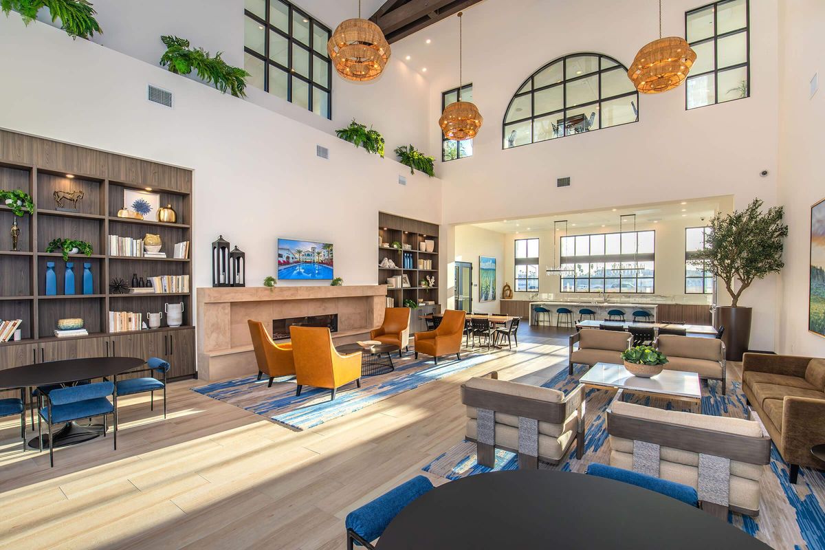 A spacious, modern interior featuring high ceilings, large windows, and stylish furnishings. There are light wood floors, comfortable seating areas with orange and beige chairs, a fireplace, bookshelves filled with decor, and hanging pendant lights, creating a warm and inviting atmosphere.