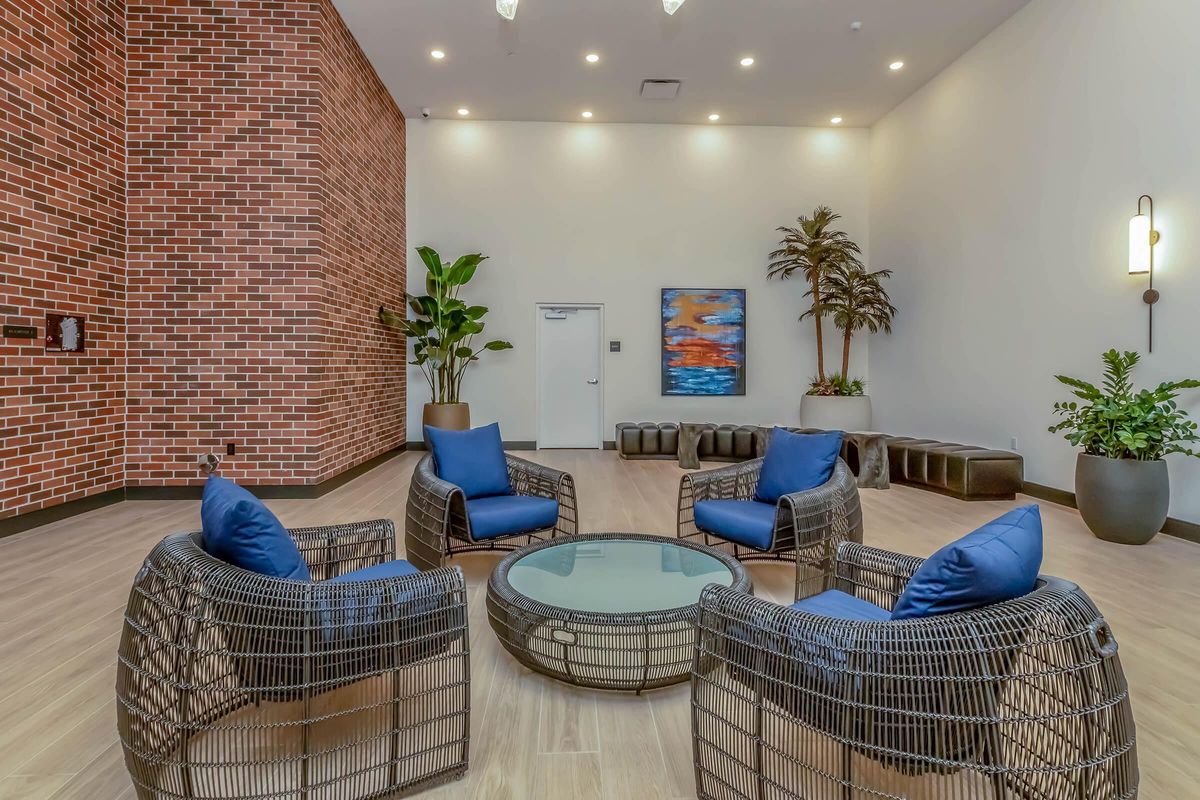 A modern indoor lounge area featuring four woven chairs with blue cushions arranged around a round glass table. The space has a brick wall, potted plants, and a piece of abstract art. Soft lighting enhances the inviting atmosphere, creating a cozy gathering spot.