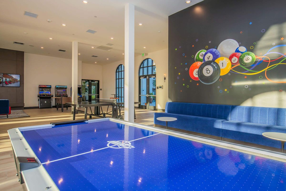Modern lounge area featuring a blue air hockey table in the foreground, a stylish blue couch, and a colorful wall mural of billiard balls. There are game machines in the background, large windows allowing natural light, and a clean, contemporary design with wooden flooring and light walls.