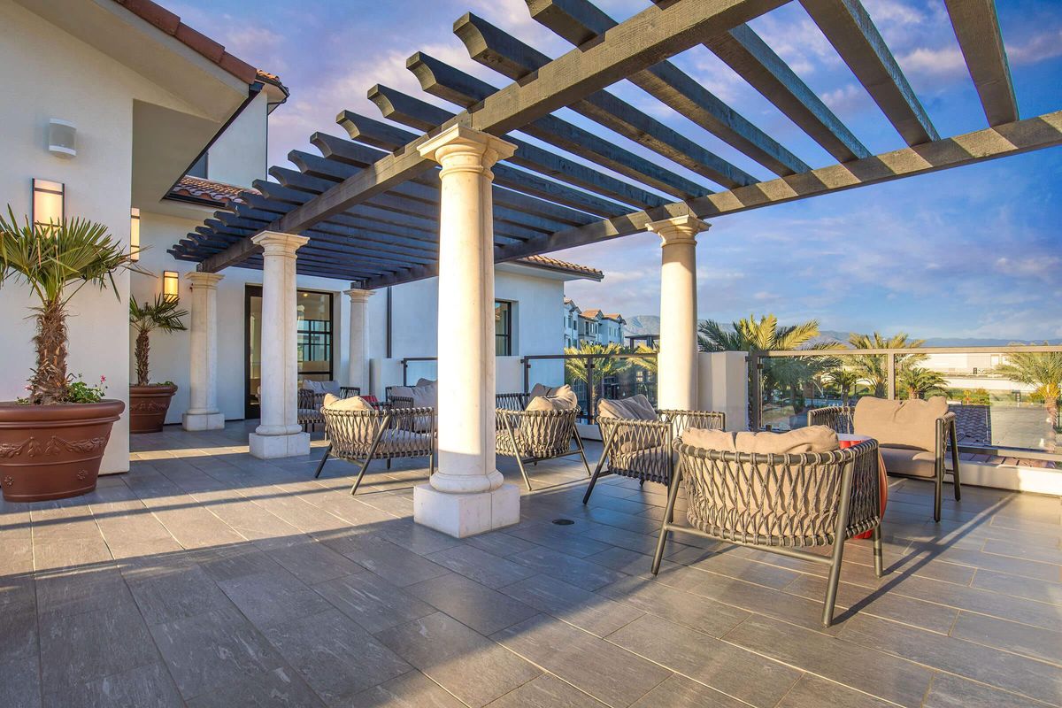 A stylish outdoor terrace featuring a pergola with black slats, surrounded by palm trees. Comfortable seating areas with woven chairs are arranged on a stone-tiled floor, offering a tranquil space with views of the sky and landscape. Ideal for relaxation or social gatherings.