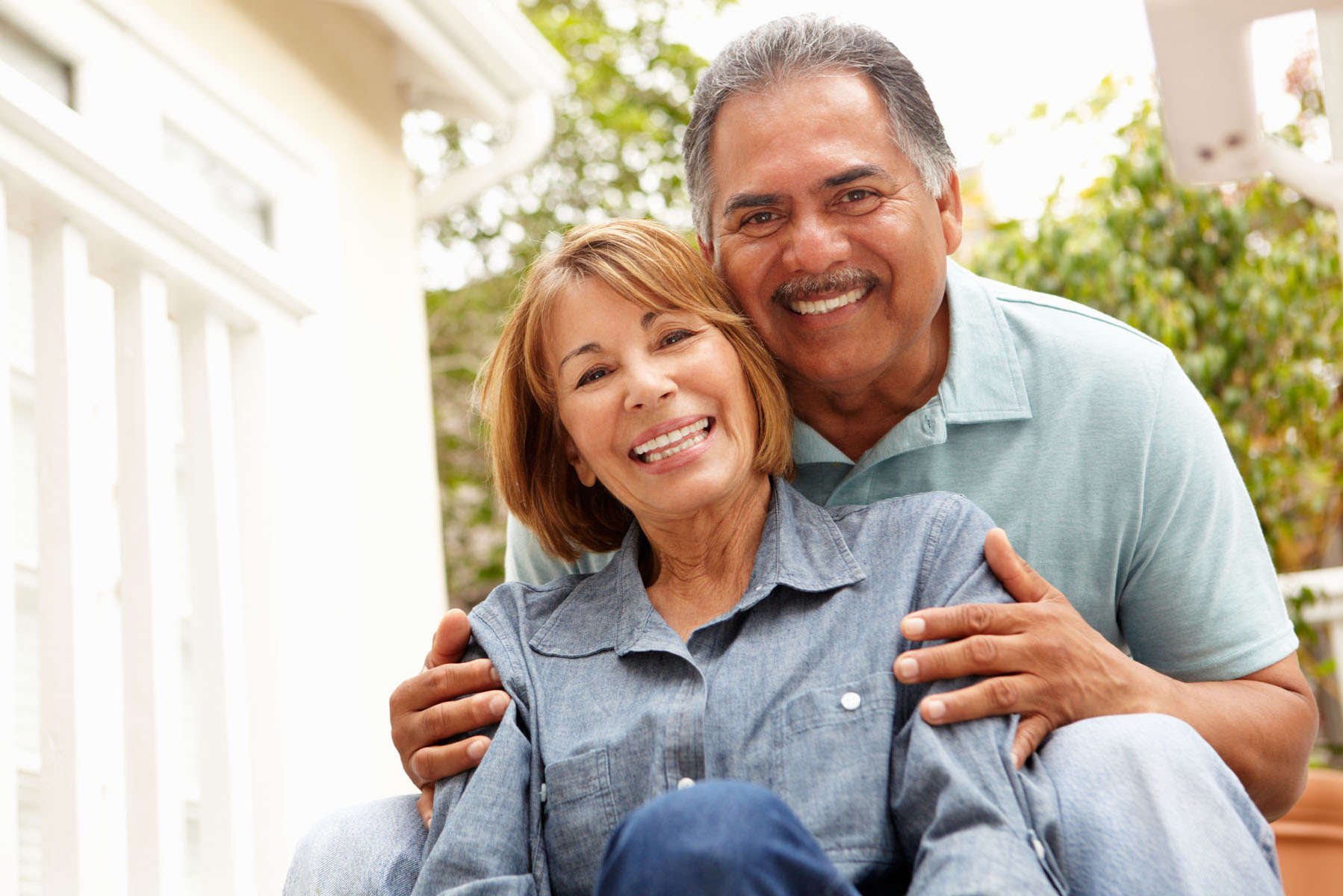 A close-up of a smiling senior couple.  The man and woman are wearing light blue shirts and are seated.  The woman is in front of the man, and the man's hands are embracing her shoulders.  Out-of-focus bushes are visible in the background.