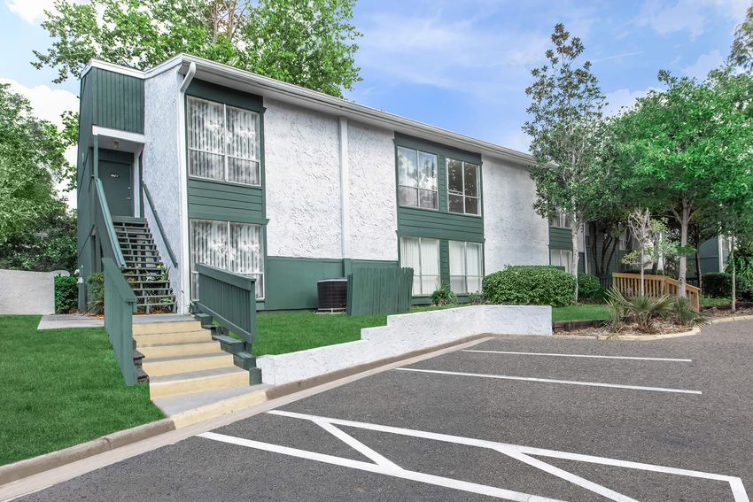 Two-story apartment building with green accents, surrounded by lush greenery. A staircase leads to the second floor, and there are several windows with curtains. The parking area in front features marked lines and well-maintained landscaping, creating a welcoming residential atmosphere.