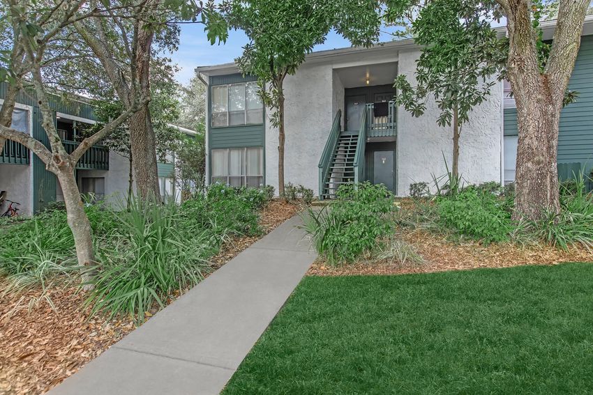 A charming apartment community featuring a two-story building with green siding. The entrance is flanked by trees and lush landscaping, with a concrete pathway leading to the front door. The area is well-maintained, showcasing a cozy and inviting atmosphere.