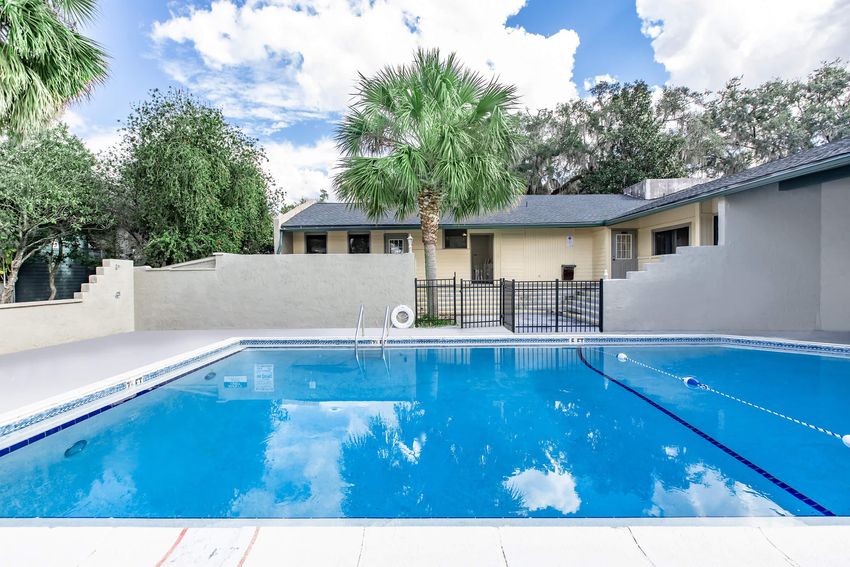 A tranquil swimming pool surrounded by a gated area, with palm trees and lush greenery in the background. The clear blue water reflects the sky and nearby structures, creating a serene atmosphere. The pool area is well-maintained with a light-colored deck.