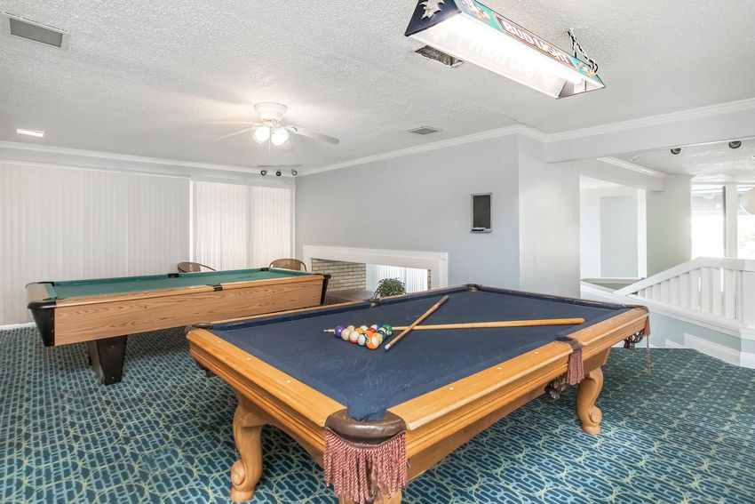 A well-lit game room featuring two pool tables. One table is set up for play with a full set of colorful billiard balls and cues laid out. The flooring is a textured blue carpet, and there are windows providing natural light. The walls are painted in light colors, creating a spacious and inviting atmosphere.